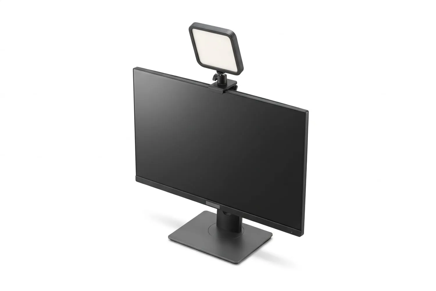 A small square LED panel clipped to the top of a monitor, lit and angled slightly down toward an empty chair, the rest of the room dim